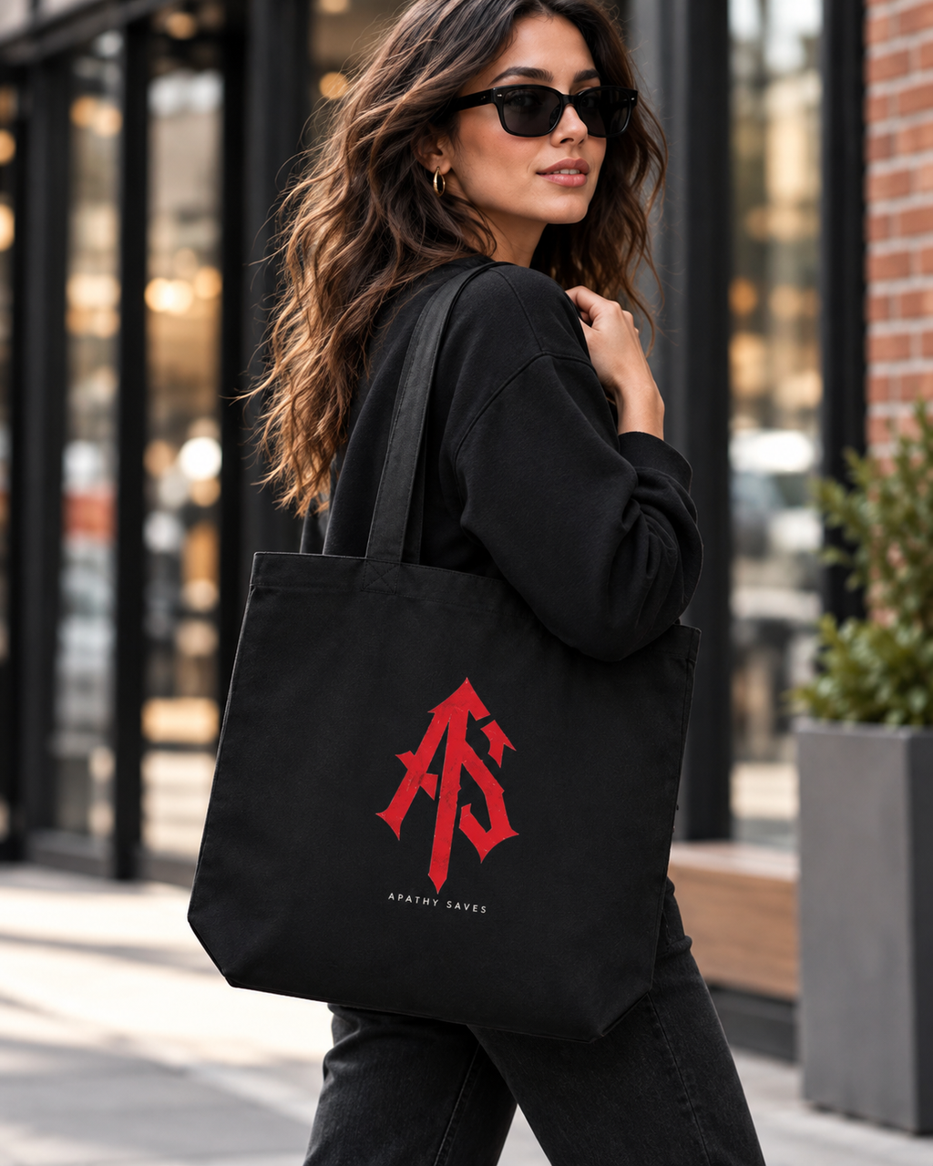 Styled lifestyle mockup of woman in black streetwear carrying ApathySaves black eco tote with centered red AS monogram and small APATHY SAVES lettering