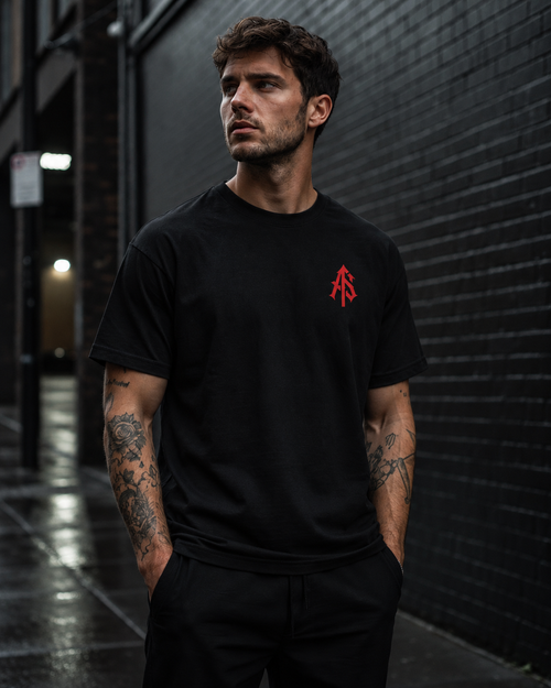 Man wearing ApathySaves black heavyweight graphic T-shirt with red interlocking AS chest logo in an urban street setting